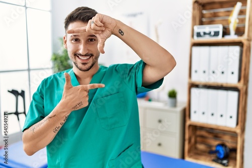 Young physiotherapist man working at pain recovery clinic smiling making frame with hands and fingers with happy face. creativity and photography concept.