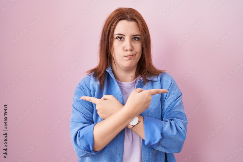 Young hispanic woman with red hair standing over pink background pointing to both sides with fingers, different direction disagree Young hispanic woman with red hair standing over pink background pointing to both sides with fingers, different direction disagree