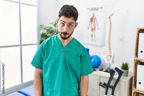Young man with beard working at pain recovery clinic making fish face with lips  crazy and comical gesture. funny expression.