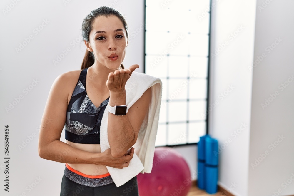 Young brunette woman wearing sportswear and towel at the gym looking at the camera blowing a kiss with hand on air being lovely and sexy. love expression.