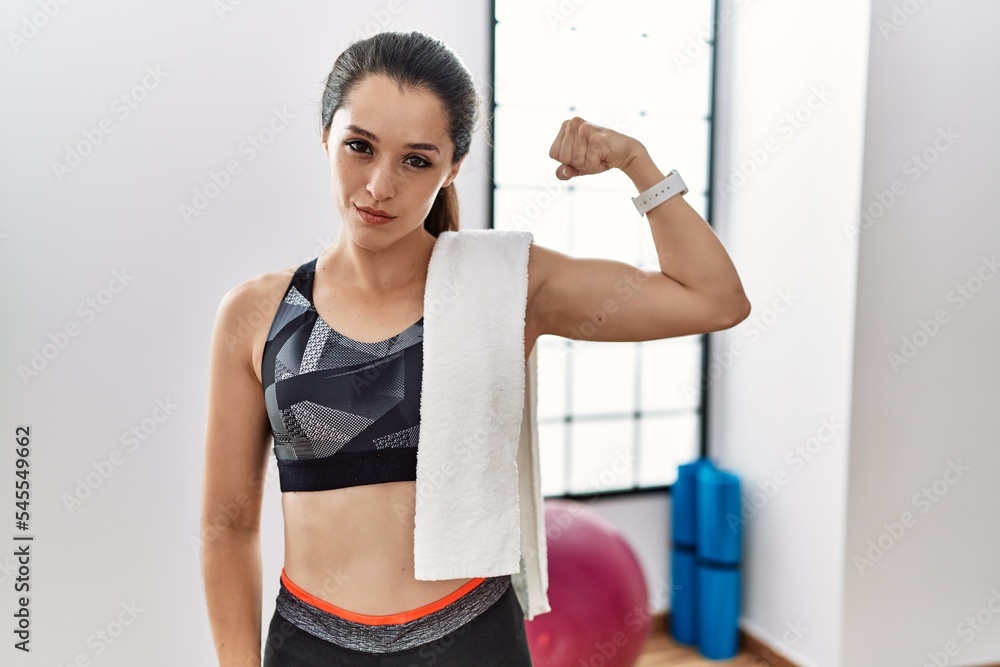 Young brunette woman wearing sportswear and towel at the gym strong person showing arm muscle, confident and proud of power