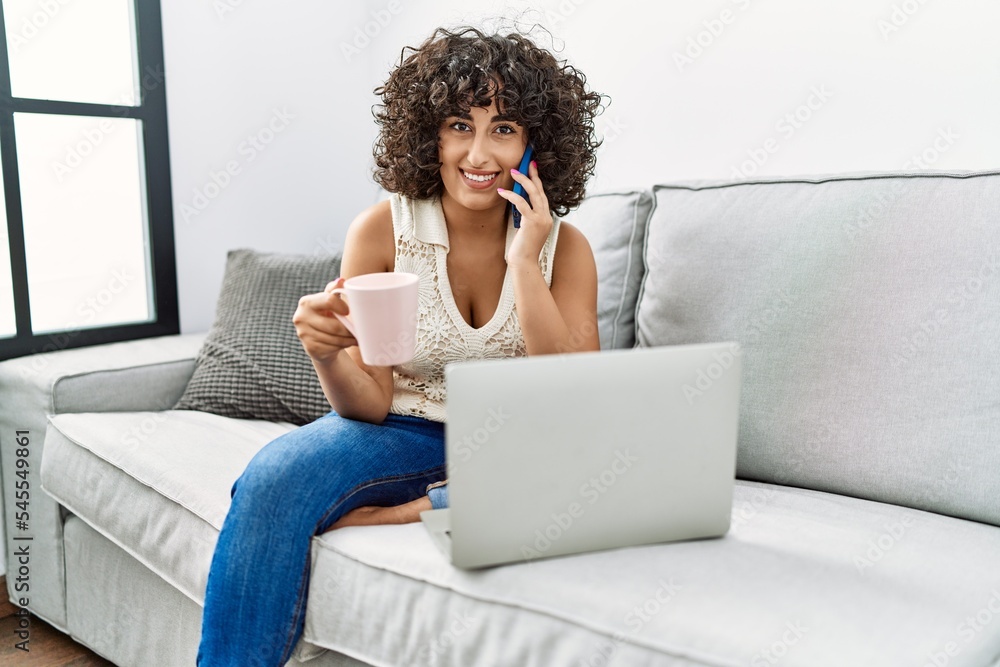 Young middle east woman drinking coffee talking on the smartphone using laptop at home Young middle east woman drinking coffee talking on the smartphone using laptop at home