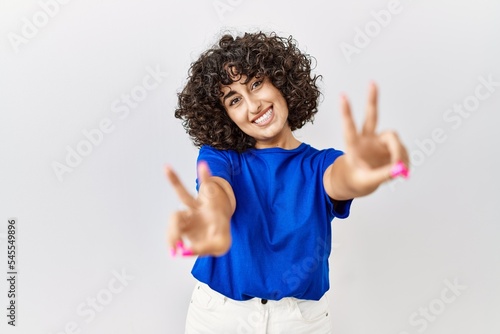 Young middle eastern woman standing over isolated background smiling looking to the camera showing fingers doing victory sign. number two.
