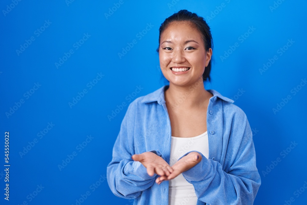 Asian young woman standing over blue background smiling with hands palms together receiving or giving gesture. hold and protection Asian young woman standing over blue background smiling with hands palms together receiving or giving gesture. hold and protection