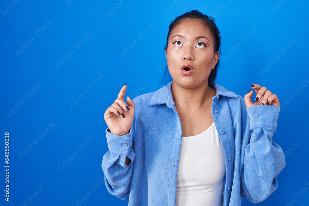 Asian young woman standing over blue background amazed and surprised looking up and pointing with fingers and raised arms. Asian young woman standing over blue background amazed and surprised looking up and pointing with fingers and raised arms.
