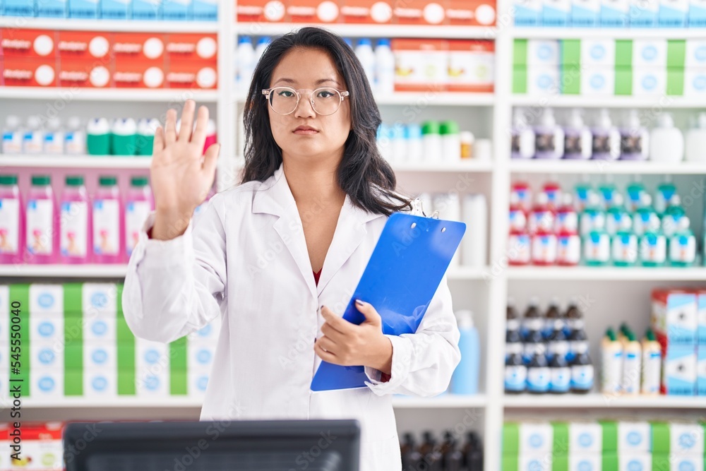 Asian young woman working at pharmacy drugstore holding clipboard with open hand doing stop sign with serious and confident expression, defense gesture