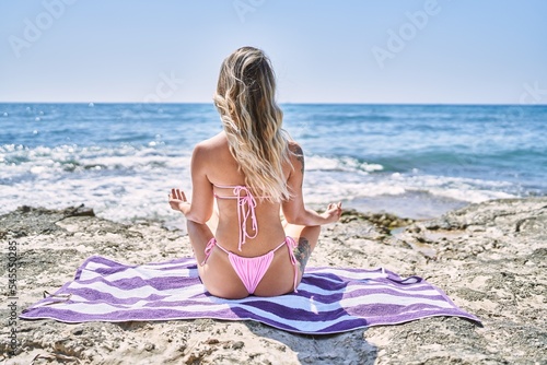 Young blonde girl on back view training yoga sitting on the towel at the beach.
