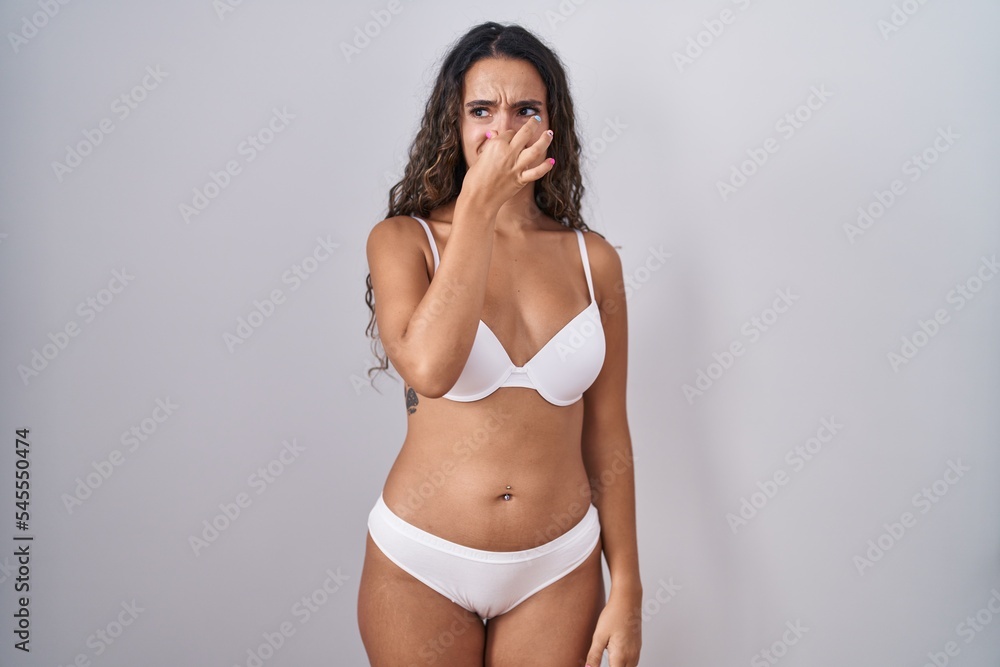 Fototapeta premium Young hispanic woman wearing white lingerie smelling something stinky and disgusting, intolerable smell, holding breath with fingers on nose. bad smell