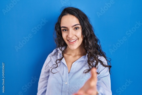 Young brunette woman standing over blue background smiling friendly offering handshake as greeting and welcoming. successful business.