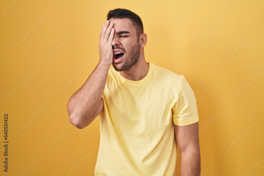 Young hispanic man standing over yellow background yawning tired covering half face, eye and mouth with hand. face hurts in pain. Young hispanic man standing over yellow background yawning tired covering half face, eye and mouth with hand. face hurts in pain.