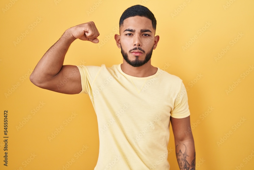 Young hispanic man standing over yellow background strong person showing arm muscle, confident and proud of power Young hispanic man standing over yellow background strong person showing arm muscle, confident and proud of power