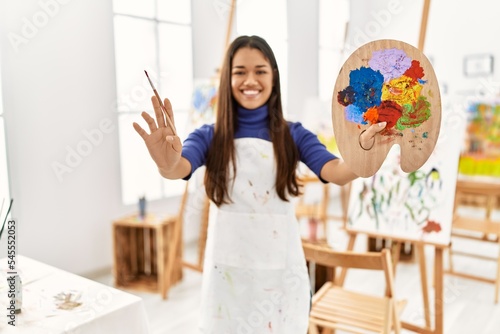 Young latin woman smiling confident holding paintbrush and palette at art studio