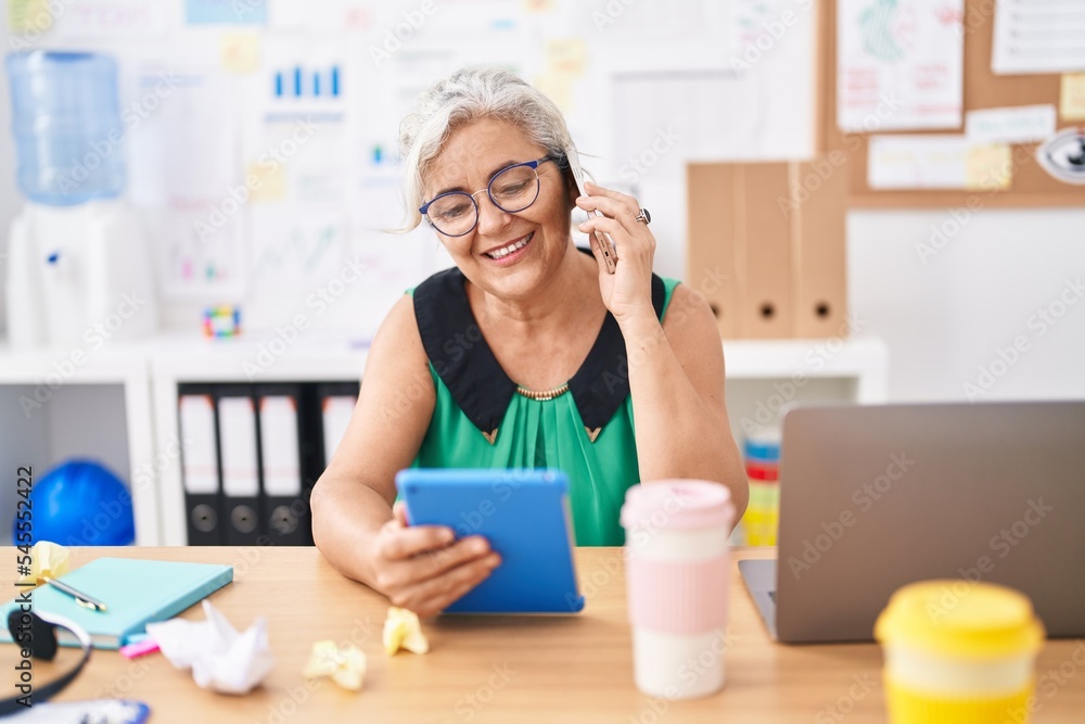 Middle age grey-haired woman business worker talking on smartphone using touchpad at office Middle age grey-haired woman business worker talking on smartphone using touchpad at office