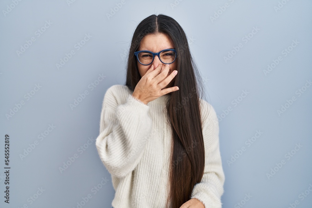 Young hispanic woman wearing casual sweater over blue background smelling something stinky and disgusting, intolerable smell, holding breath with fingers on nose. bad smell Young hispanic woman wearing casual sweater over blue background smelling something stinky and disgusting, intolerable smell, holding breath with fingers on nose. bad smell