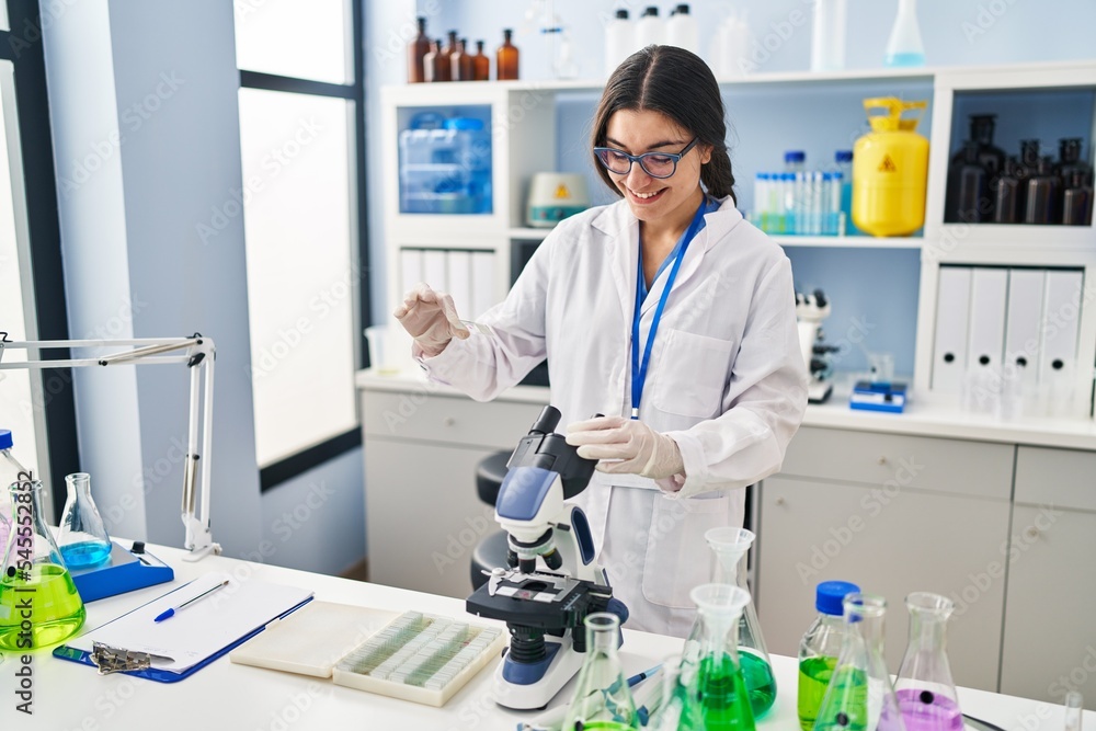 Young hispanic woman wearing scientist uniform using microscope at