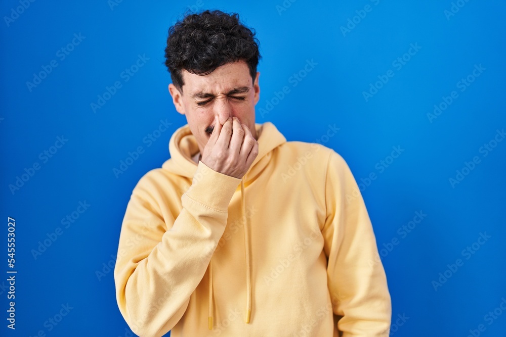 Hispanic man standing over blue background smelling something stinky and disgusting, intolerable smell, holding breath with fingers on nose. bad smell