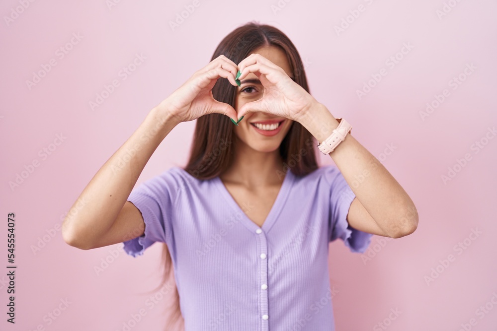 Young hispanic woman with long hair standing over pink background doing heart shape with hand and fingers smiling looking through sign Young hispanic woman with long hair standing over pink background doing heart shape with hand and fingers smiling looking through sign
