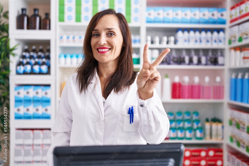 Middle age brunette woman working at pharmacy drugstore showing and pointing up with fingers number two while smiling confident and happy.