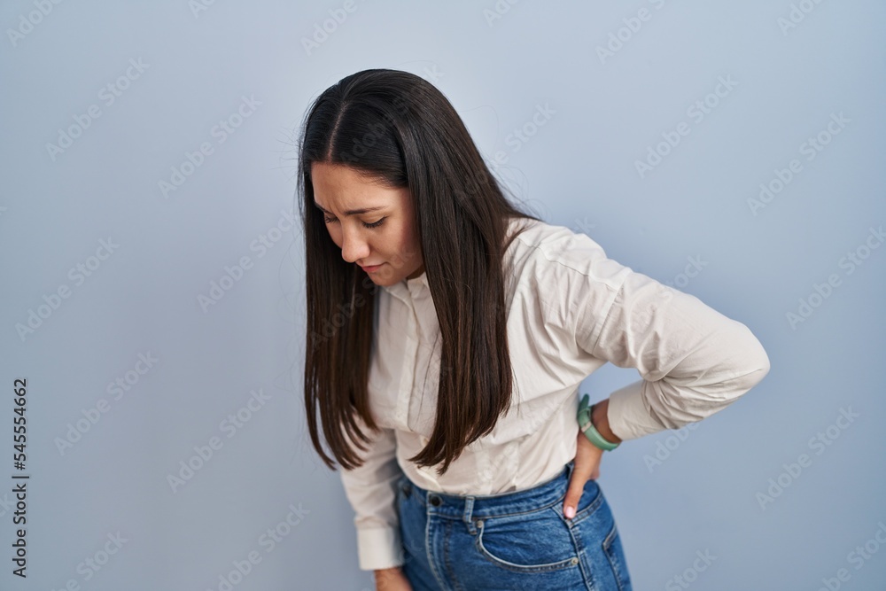 Young latin woman standing over blue background suffering of backache, touching back with hand, muscular pain Young latin woman standing over blue background suffering of backache, touching back with hand, muscular pain