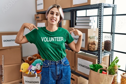 Young beautiful woman wearing volunteer t shirt at donations stand looking confident with smile on face, pointing oneself with fingers proud and happy.