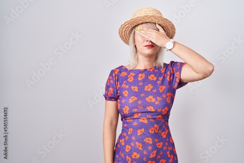 Young caucasian woman wearing flowers dress and summer hat covering eyes with hand, looking serious and sad. sightless, hiding and rejection concept