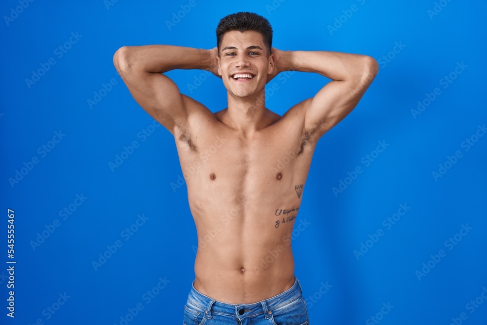 Young hispanic man standing shirtless over blue background relaxing and stretching, arms and hands behind head and neck smiling happy