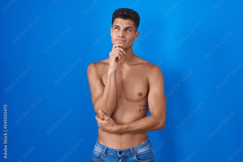 Young hispanic man standing shirtless over blue background with hand on chin thinking about question, pensive expression. smiling with thoughtful face. doubt concept.