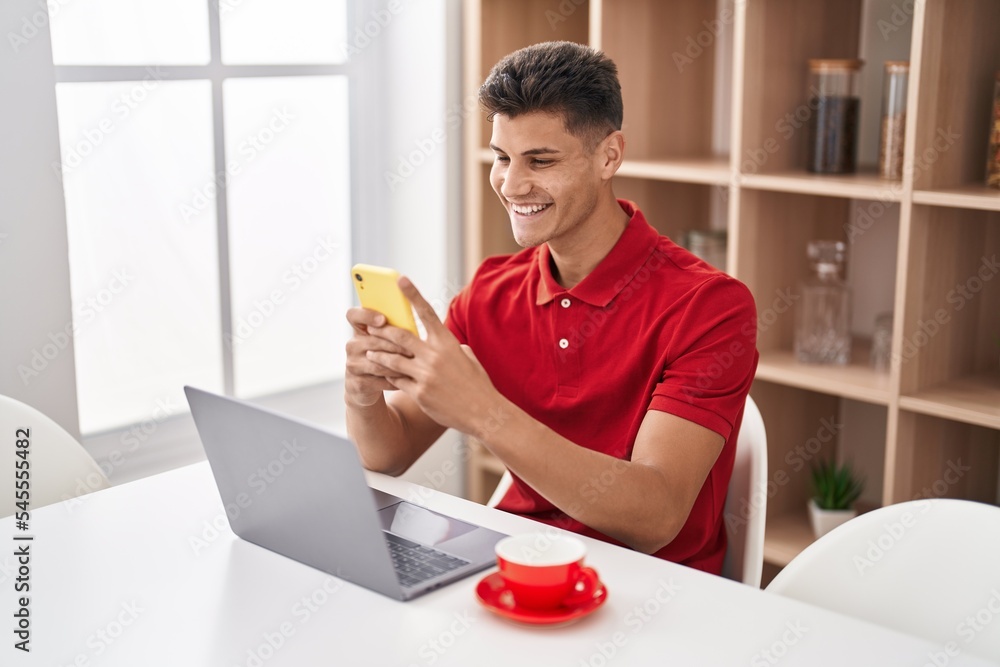 Young hispanic man using laptop and smartphone sitting on table at home Young hispanic man using laptop and smartphone sitting on table at home