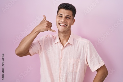 Young hispanic man standing over pink background smiling doing phone gesture with hand and fingers like talking on the telephone. communicating concepts.