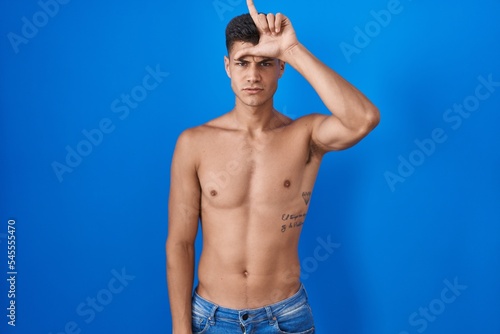 Young hispanic man standing shirtless over blue background making fun of people with fingers on forehead doing loser gesture mocking and insulting.