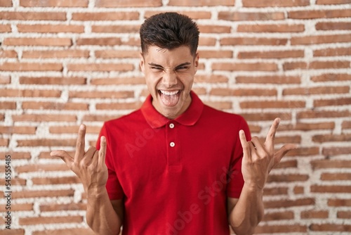 Young hispanic man standing over bricks wall shouting with crazy expression doing rock symbol with hands up. music star. heavy concept.