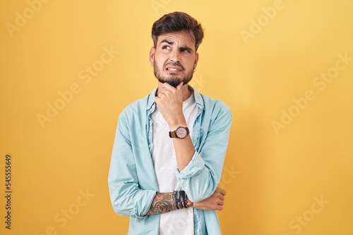 Young hispanic man with tattoos standing over yellow background thinking worried about a question  concerned and nervous with hand on chin