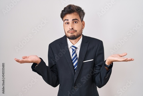 Young hispanic man with tattoos wearing business suit and tie clueless and confused expression with arms and hands raised. doubt concept.