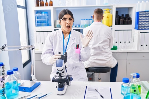Young woman working at scientist laboratory scared and amazed with open mouth for surprise  disbelief face