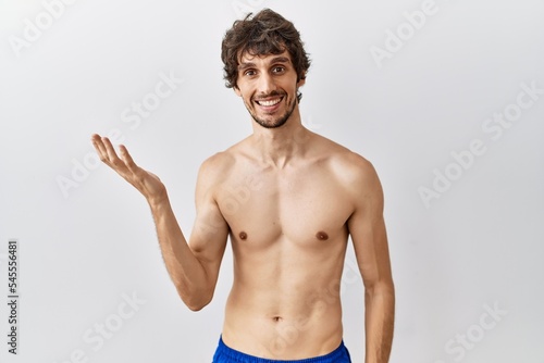 Young hispanic man standing shirtless over isolated, background smiling cheerful presenting and pointing with palm of hand looking at the camera.