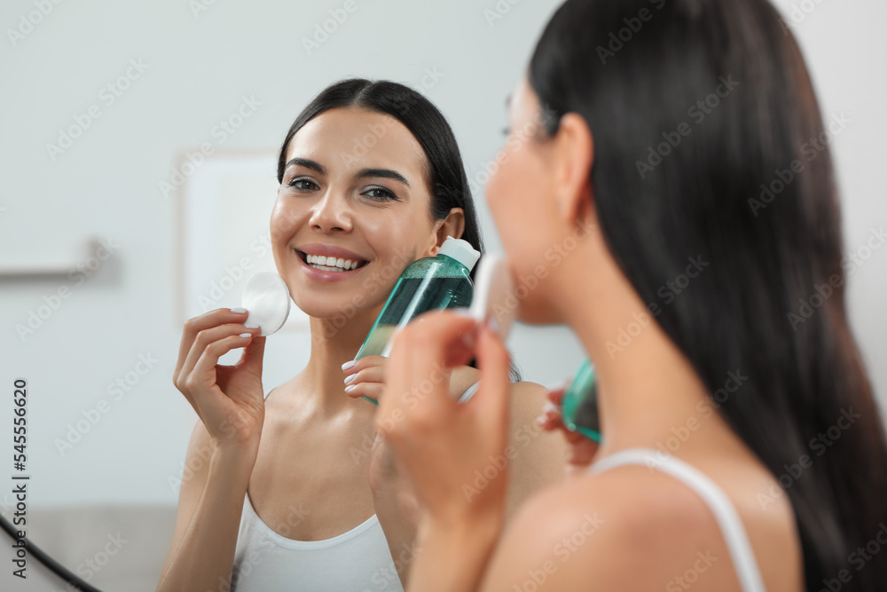 Young woman using cotton pad with micellar water near mirror indoors Young woman using cotton pad with micellar water near mirror indoors