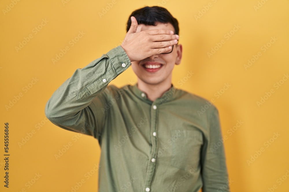Non binary person standing over yellow background smiling and laughing with hand on face covering eyes for surprise. blind concept.