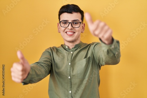 Non binary person standing over yellow background approving doing positive gesture with hand, thumbs up smiling and happy for success. winner gesture.