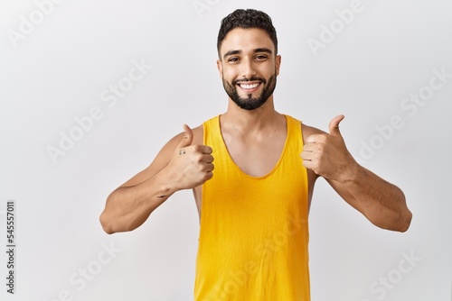Young handsome man with beard standing over isolated background success sign doing positive gesture with hand, thumbs up smiling and happy. cheerful expression and winner gesture.