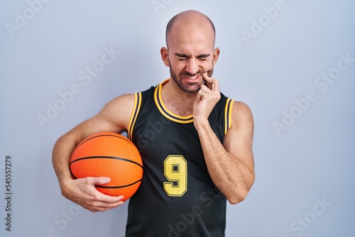 Young bald man with beard wearing basketball uniform holding ball touching mouth with hand with painful expression because of toothache or dental illness on teeth. dentist concept.