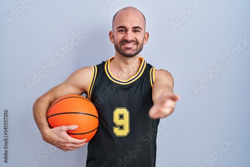 Young bald man with beard wearing basketball uniform holding ball smiling friendly offering handshake as greeting and welcoming. successful business.