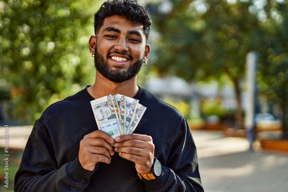 Young arab man smiling confident holding sol peruvian banknotes at park - obrazy, fototapety, plakaty 