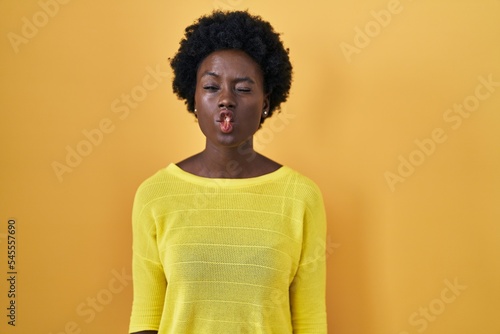 African young woman standing over yellow studio making fish face with lips, crazy and comical gesture. funny expression.