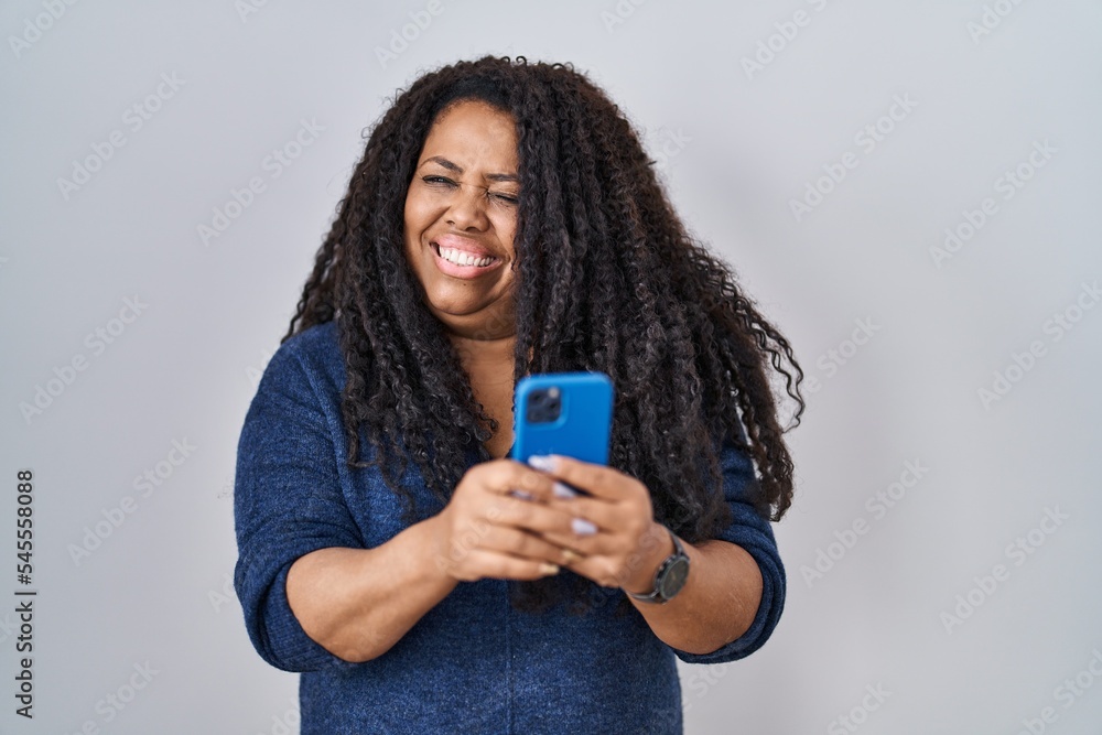 Plus size hispanic woman using smartphone typing message winking looking at the camera with sexy expression, cheerful and happy face. Plus size hispanic woman using smartphone typing message winking looking at the camera with sexy expression, cheerful and happy face.