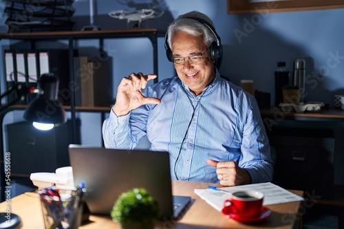 Hispanic senior man wearing call center agent headset at night smiling and confident gesturing with hand doing small size sign with fingers looking and the camera. measure concept.