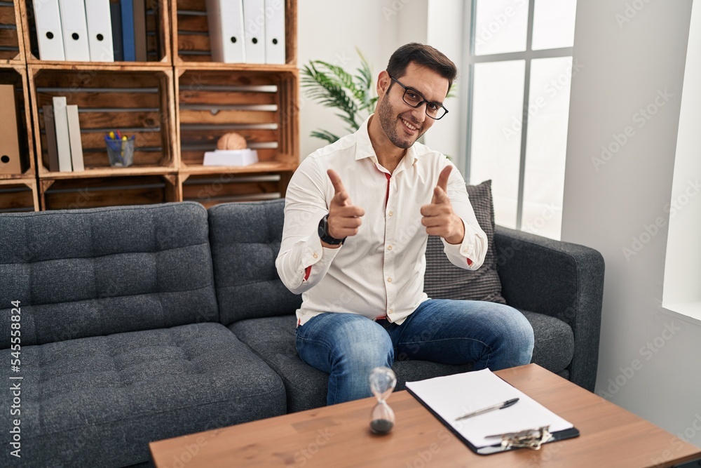 Young hispanic man with beard working at consultation office pointing fingers to camera with happy and funny face. good energy and vibes. Young hispanic man with beard working at consultation office pointing fingers to camera with happy and funny face. good energy and vibes.