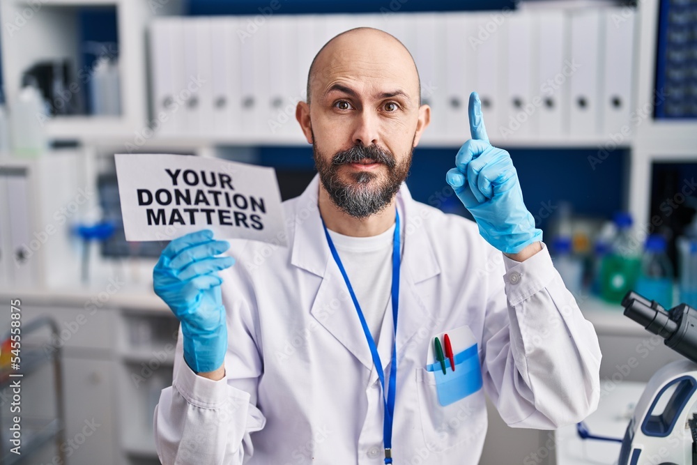 Young hispanic man working at scientist laboratory holding you donation matters banner surprised with an idea or question pointing finger with happy face, number one Young hispanic man working at scientist laboratory holding you donation matters banner surprised with an idea or question pointing finger with happy face, number one