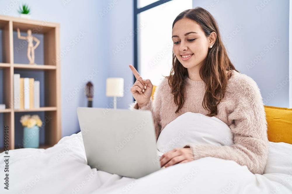 Young hispanic woman using computer laptop on the bed smiling happy pointing with hand and finger to the side Young hispanic woman using computer laptop on the bed smiling happy pointing with hand and finger to the side