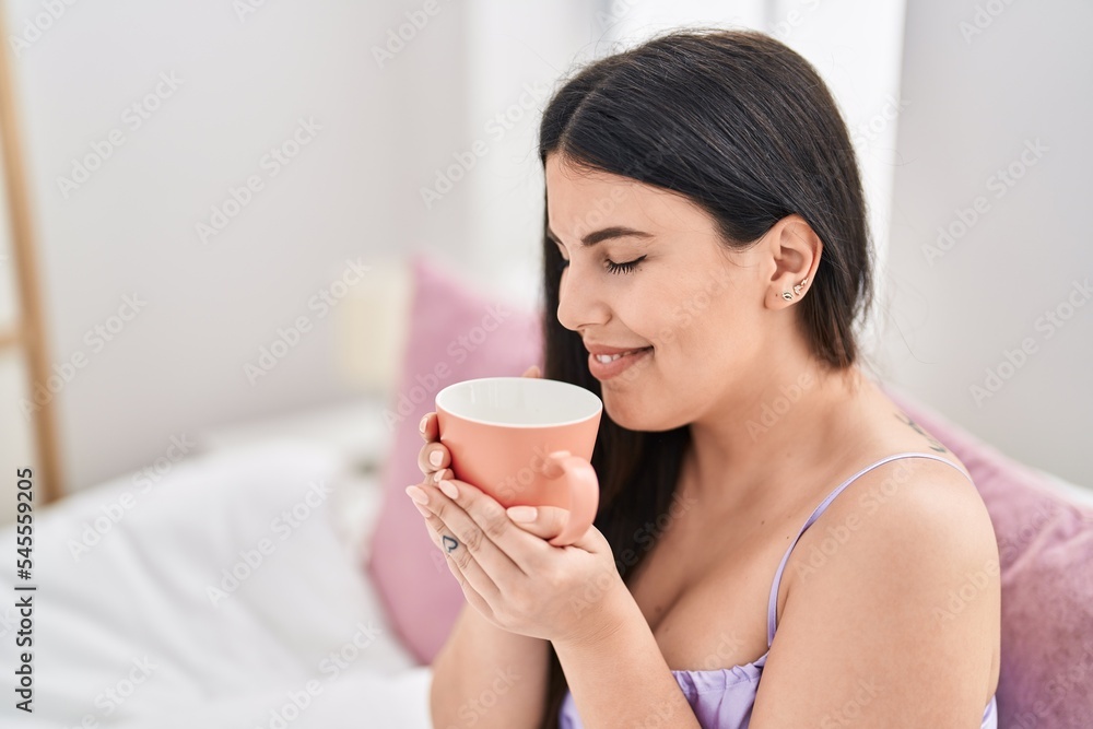 Young hispanic woman drinking cup of coffee sitting on bed at bedroom Young hispanic woman drinking cup of coffee sitting on bed at bedroom