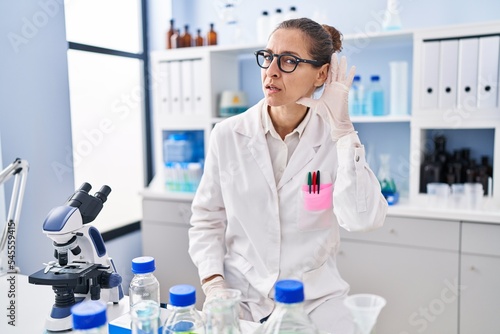 Young woman working at scientist laboratory smiling with hand over ear listening and hearing to rumor or gossip. deafness concept.